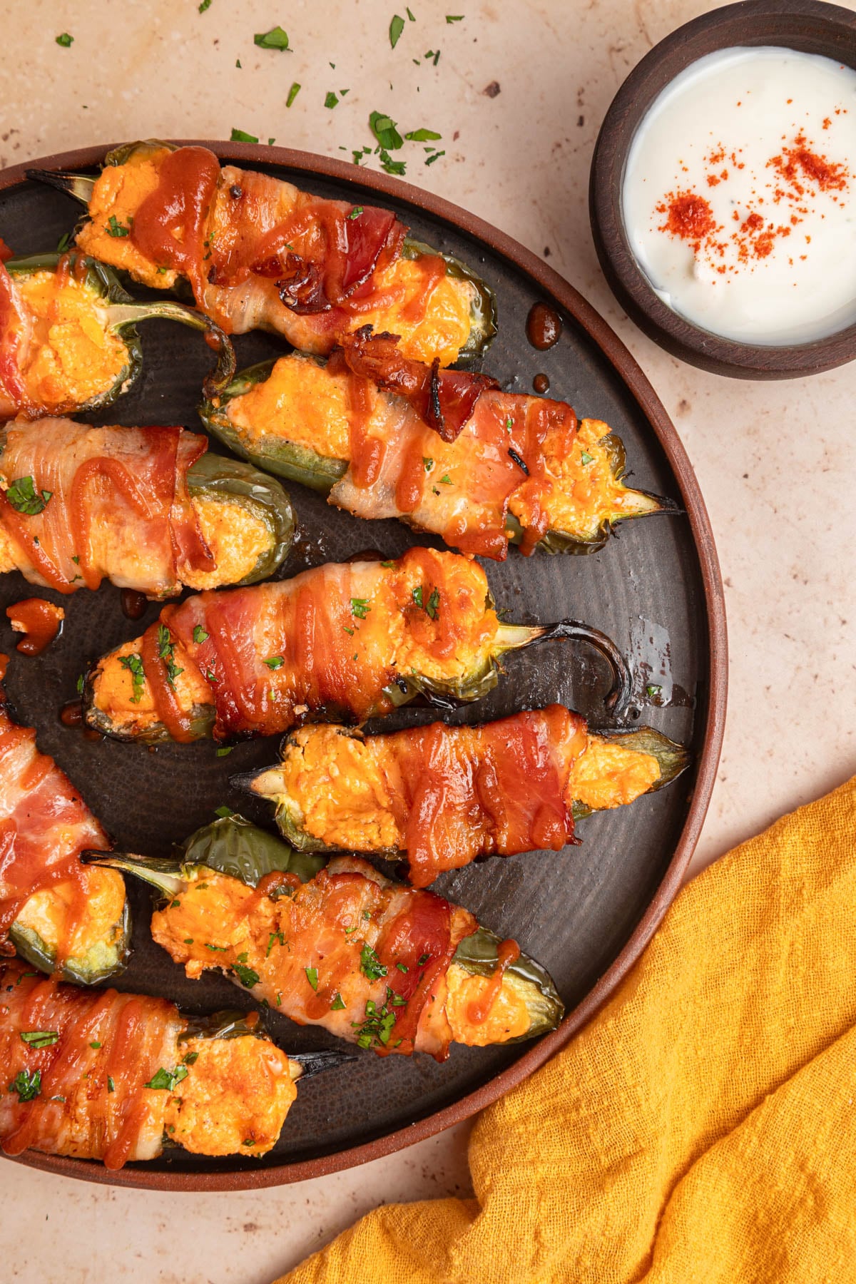 A plate of bacon-wrapped stuffed jalapeños with melted cheese, garnished with herbs, sits beside a bowl of white dipping sauce and a yellow cloth napkin.