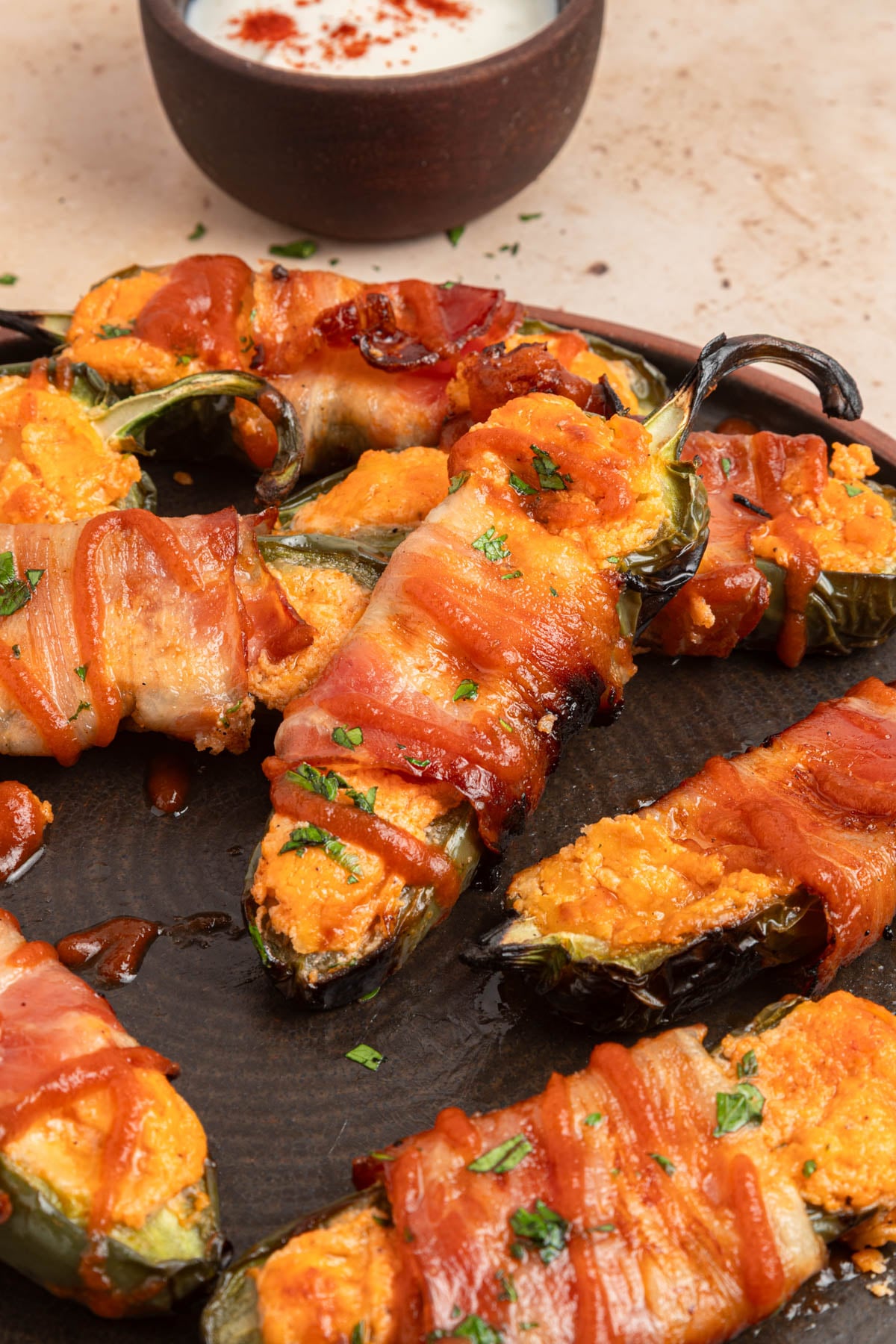 Bacon-wrapped jalapeño poppers stuffed with cheese, garnished with herbs, served on a dark plate with a bowl of dipping sauce in the background.