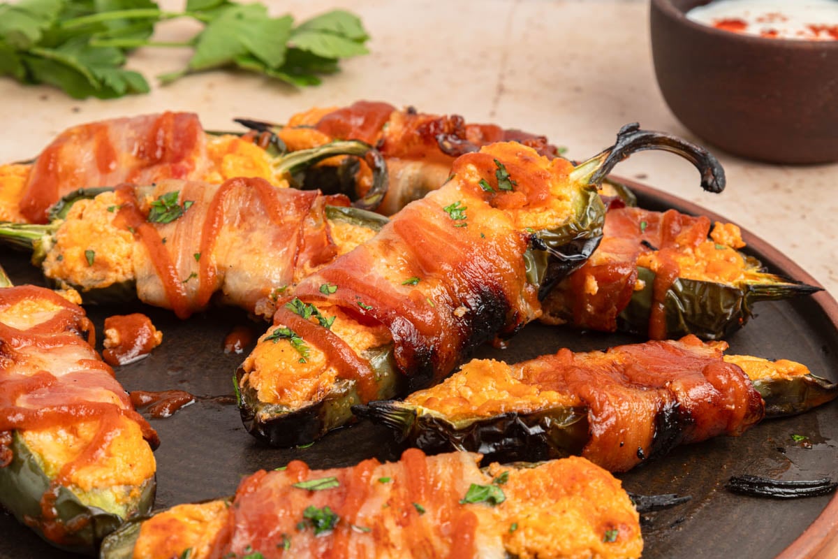 Bacon-wrapped jalapeño peppers stuffed with cheese are arranged on a dark plate, garnished with herbs, with a dip in a small bowl nearby.