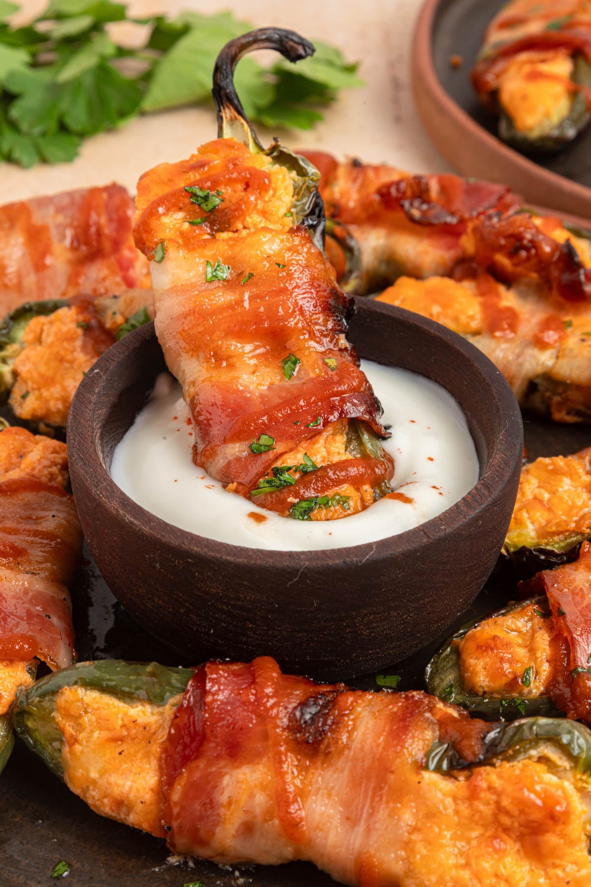Bacon-wrapped jalapeño popper being dipped into a bowl of white sauce, surrounded by more poppers on a dark plate.
