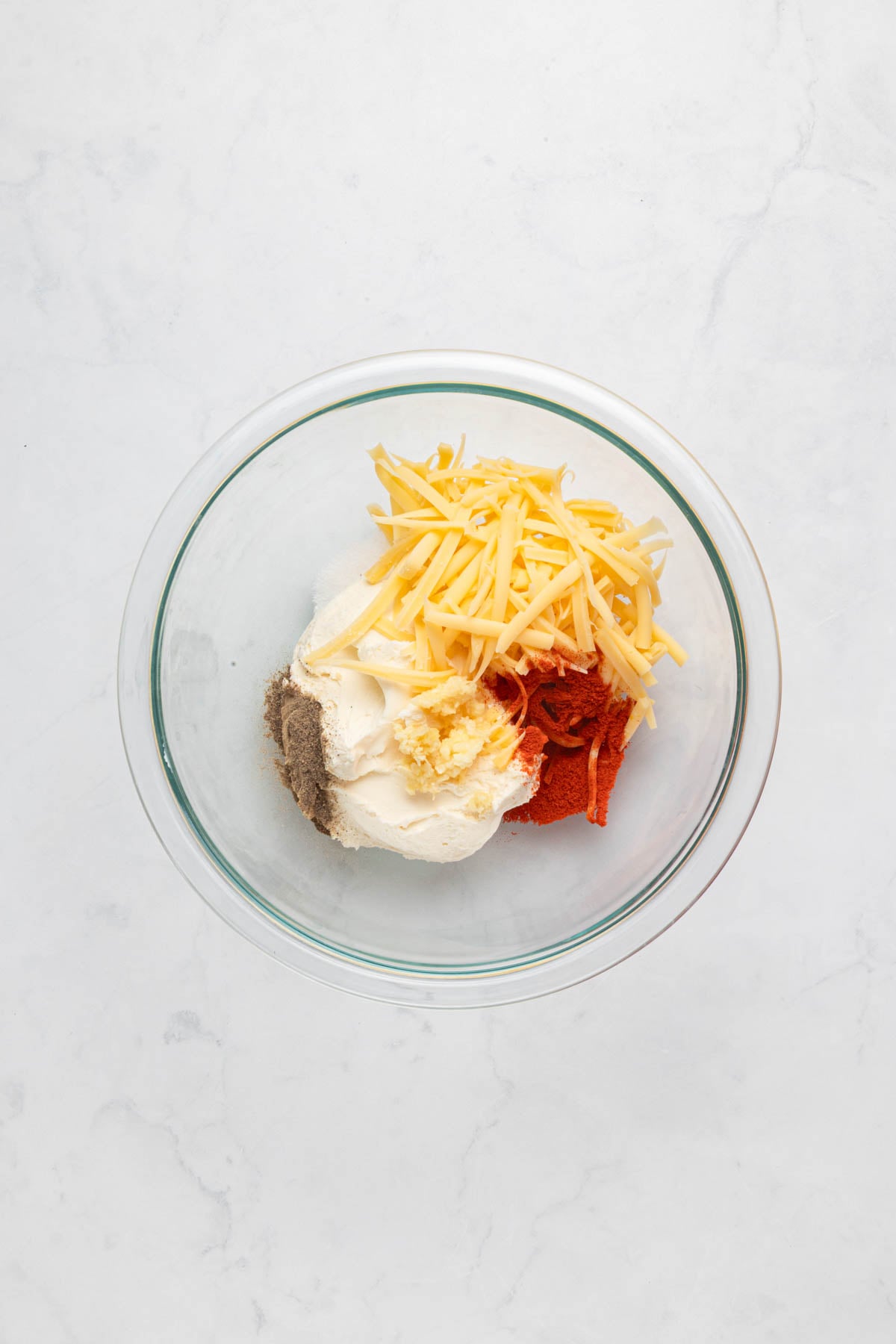 A glass bowl containing shredded cheese, cream cheese, ground black pepper, minced garlic, and paprika on a white marble surface.
