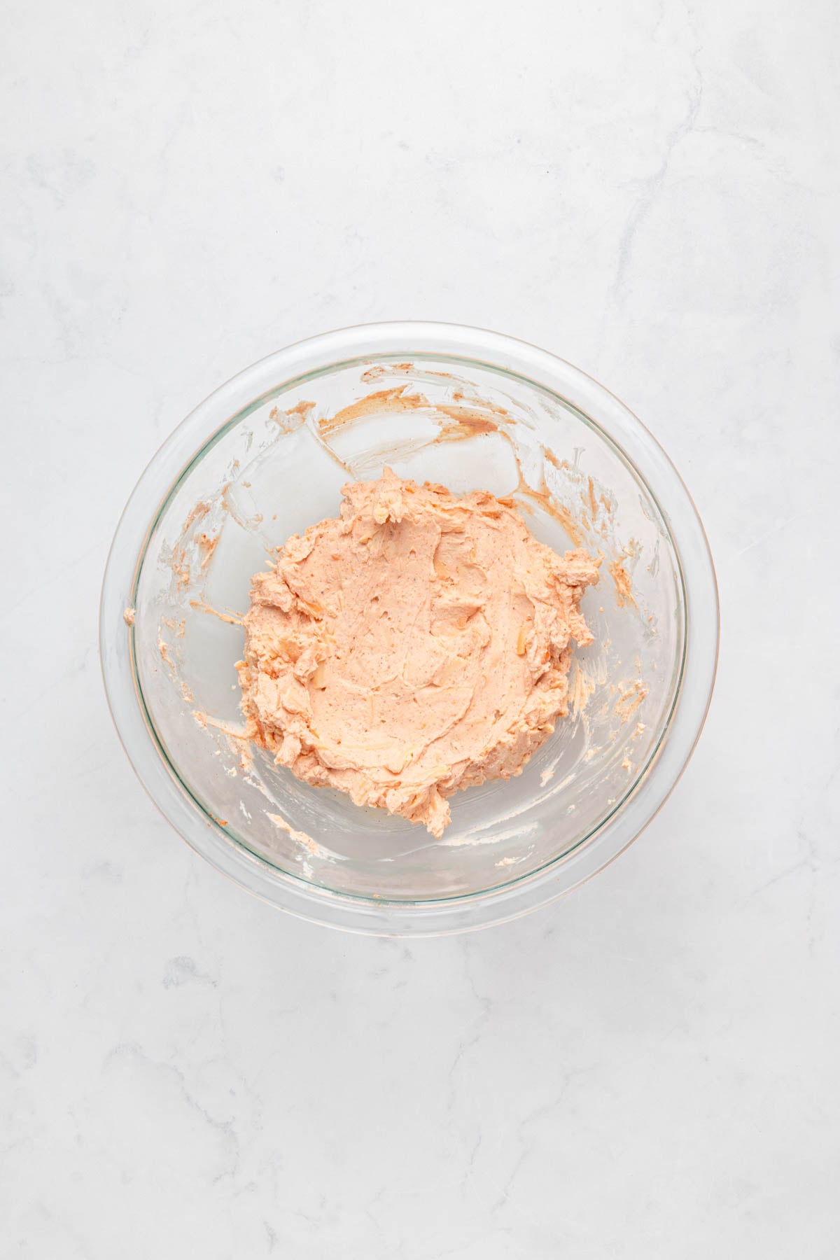 A glass bowl containing a light brown, creamy mixture sits on a white marble surface.