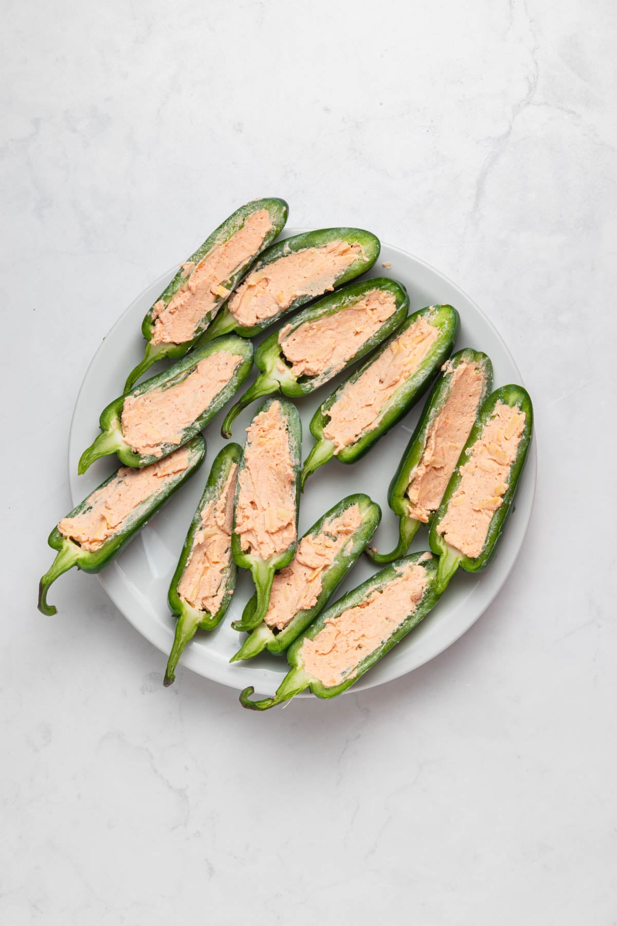 A white plate with halved jalapeños filled with a creamy, orange cheese mixture on a light, marble surface.