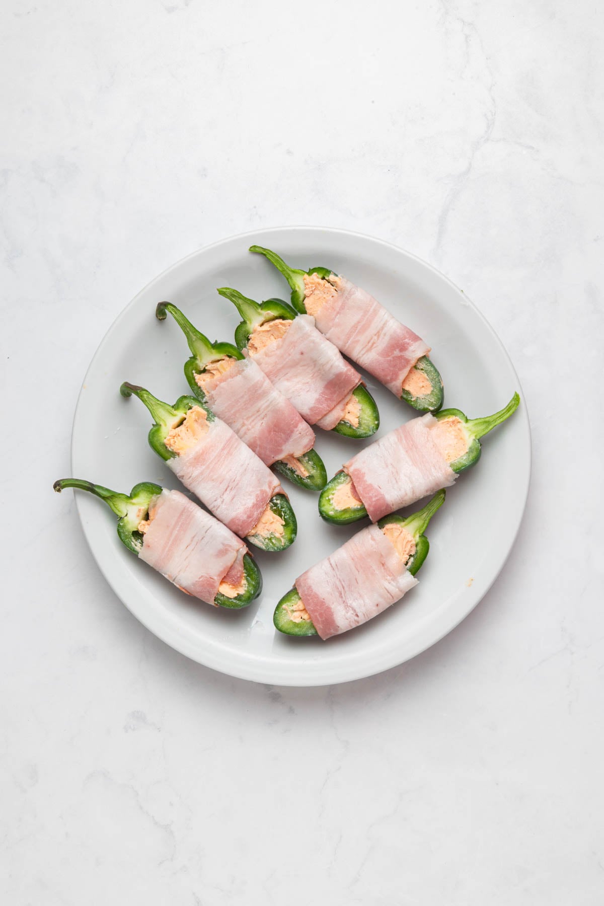A white plate with six jalapeño halves stuffed with cheese and wrapped in raw bacon, arranged on a light marble surface.