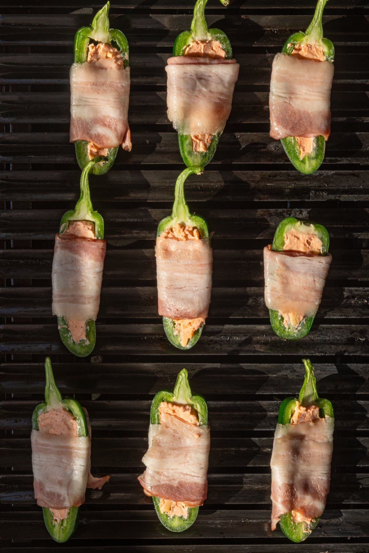 Nine jalapeño halves stuffed with cheese and wrapped in bacon are arranged in rows on a grill.