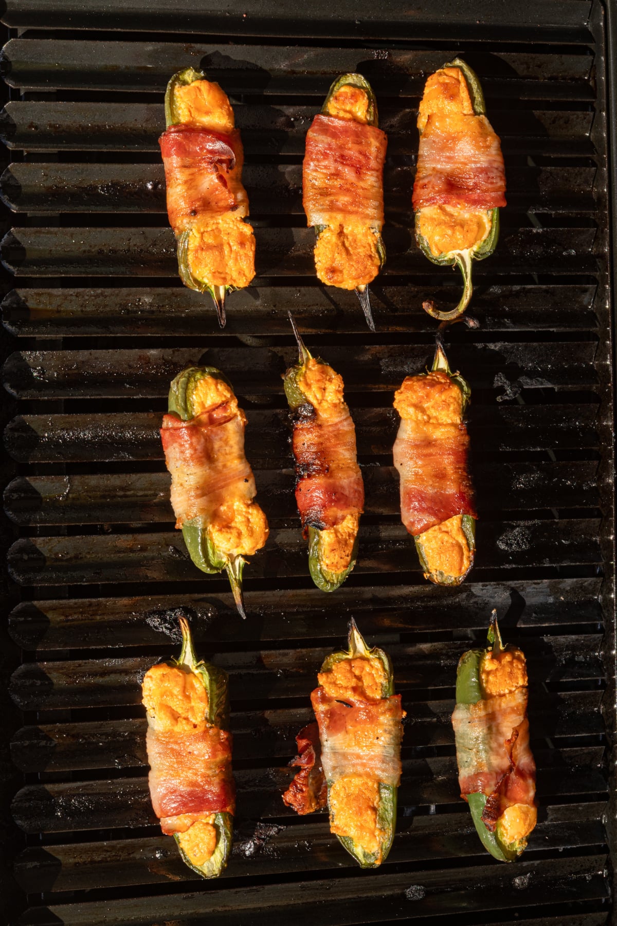Eight bacon-wrapped, cheese-stuffed jalapeño halves are grilling on a black grill grate.