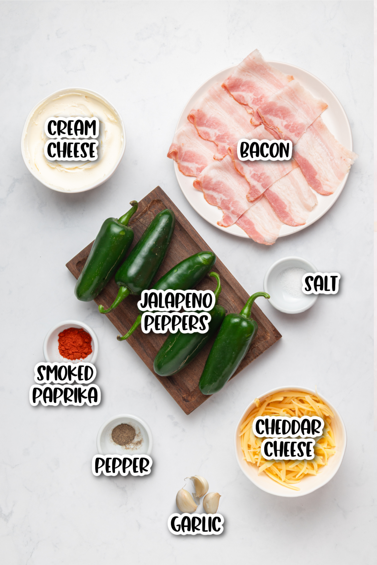 Ingredients for jalapeño poppers arranged on a white surface: cream cheese, bacon, jalapeño peppers, salt, smoked paprika, pepper, garlic, and cheddar cheese.