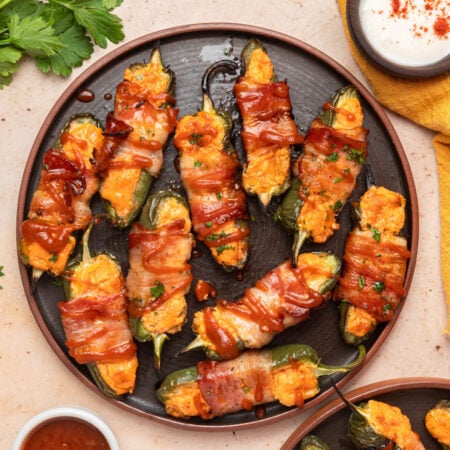 A plate of jalapeño poppers stuffed with cheese filling, wrapped in bacon, and drizzled with sauce, served alongside dips and garnished with chopped herbs.