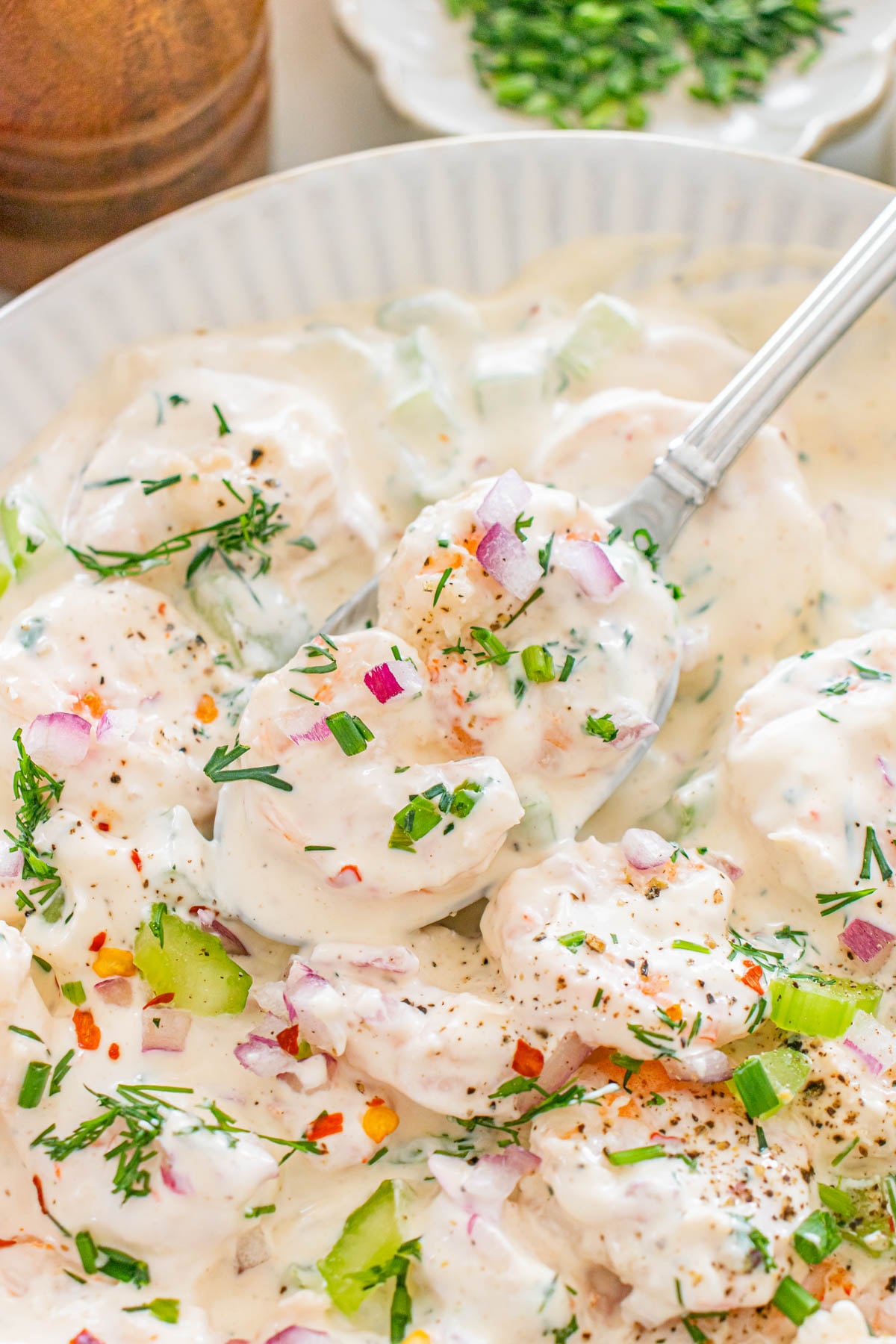 A close-up of a creamy shrimp salad garnished with chopped red onions, celery, fresh herbs, and red pepper flakes in a white bowl with a serving spoon.