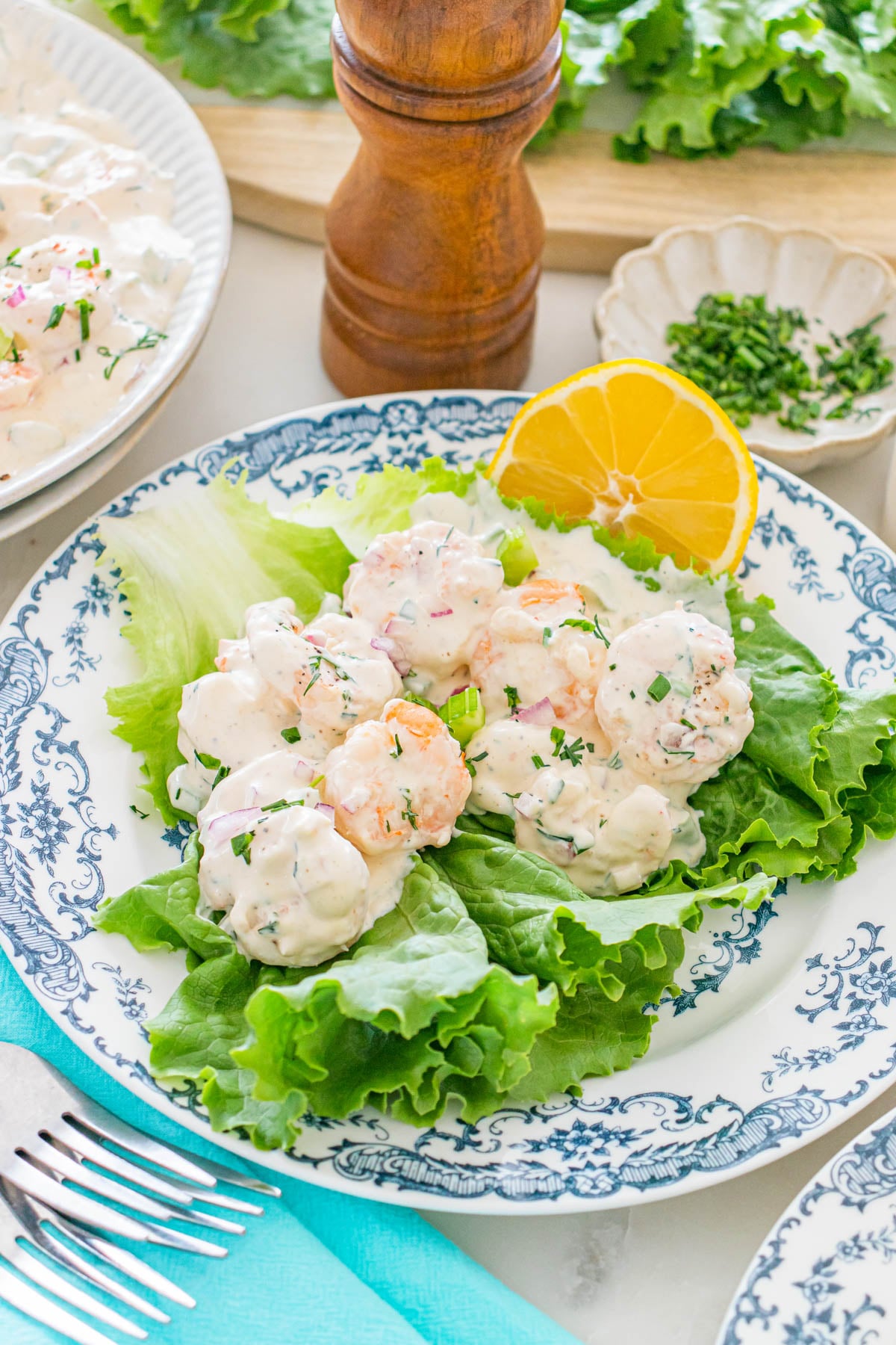 A plate with creamy shrimp salad served on lettuce leaves, garnished with a lemon slice and fresh herbs, on a decorative blue and white dish.