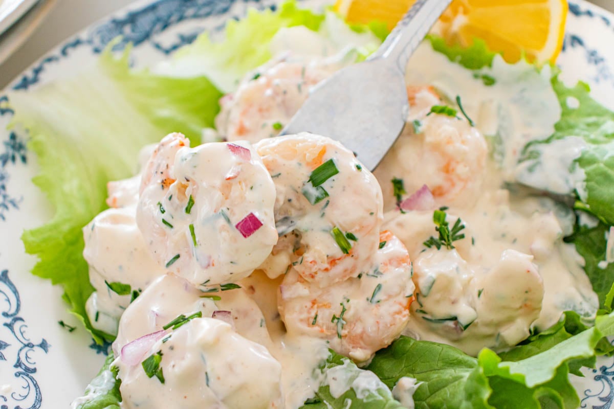 Shrimp coated in a creamy sauce with chopped herbs and red onion, served on a bed of lettuce with a slice of orange on a decorative plate.