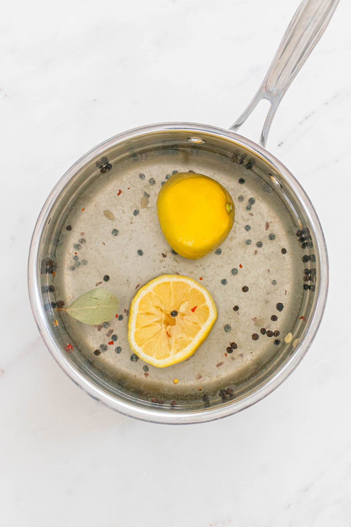 A saucepan containing water, lemon halves, a bay leaf and peppercorns on a white surface.