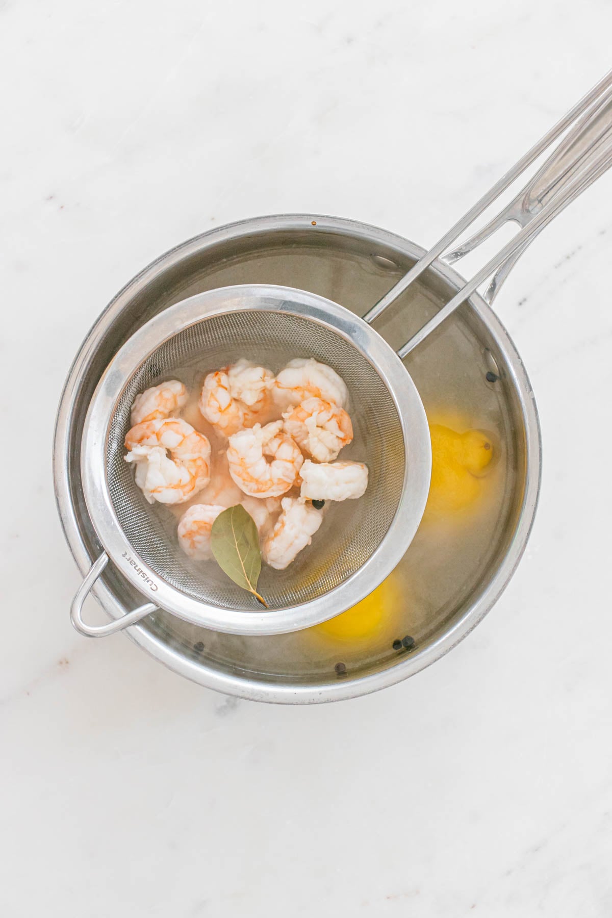 A metal strainer with cooked shrimp and a bay leaf is placed over a pot of water containing lemon halves and peppercorns.