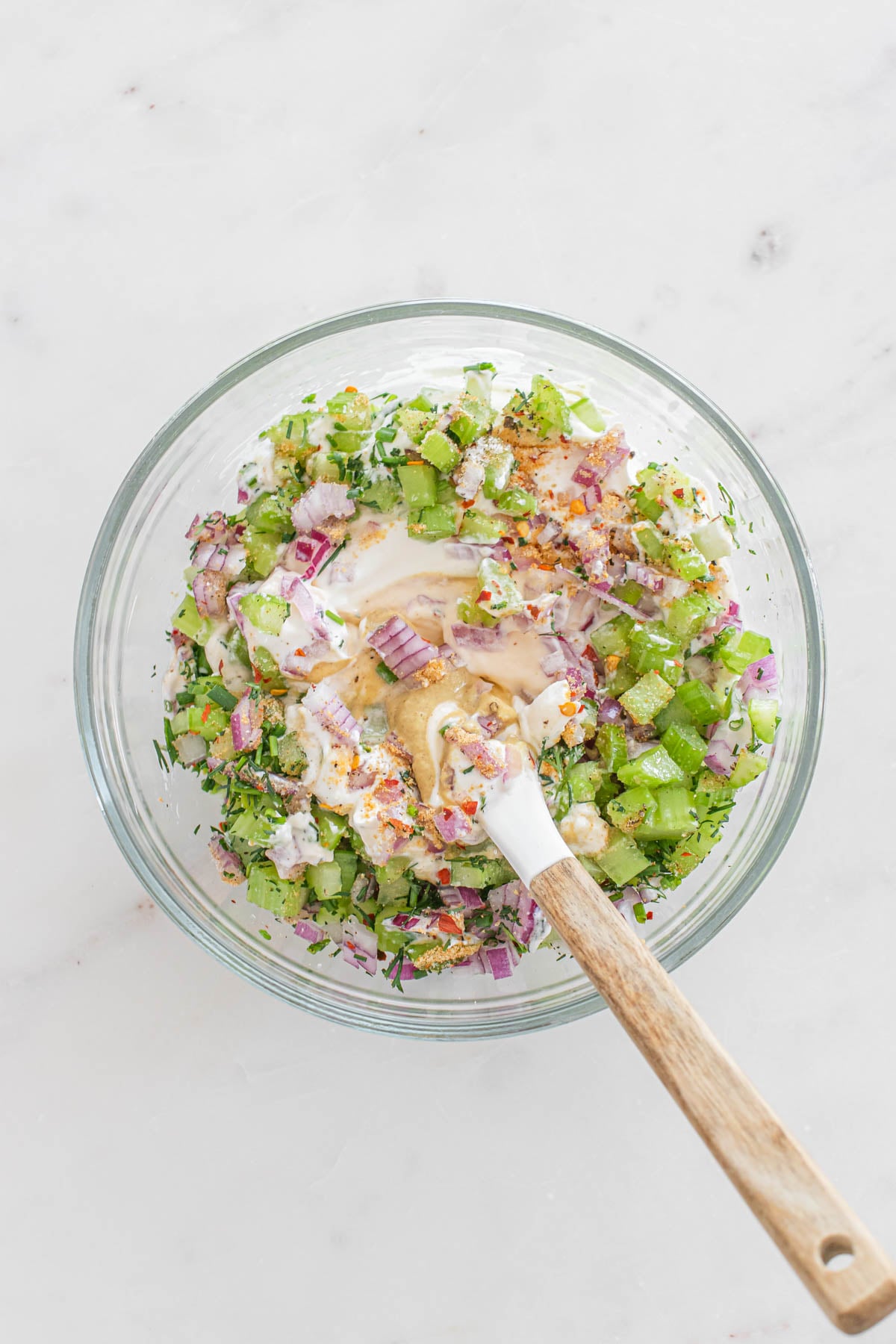 A glass bowl with chopped celery, red onion, herbs, and a creamy dressing being mixed with a wooden spoon on a white surface.