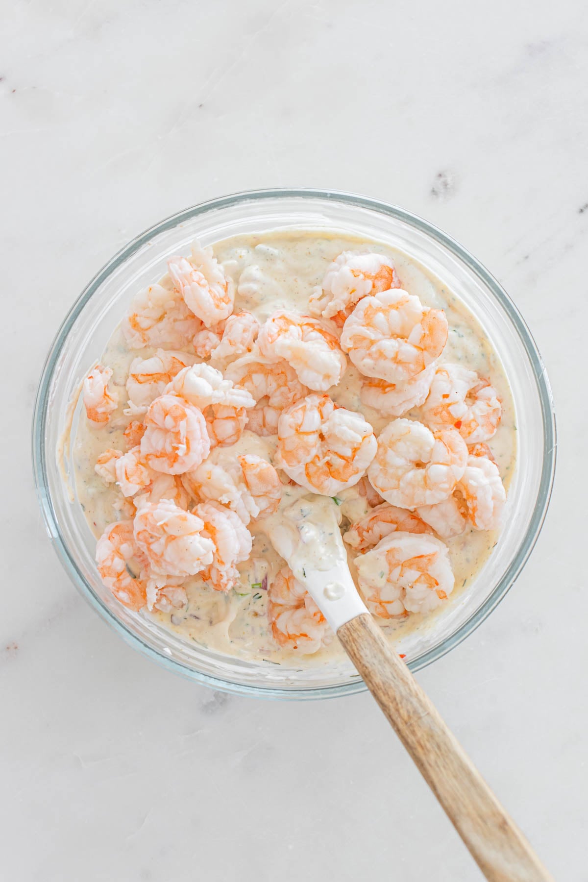 A glass bowl filled with shrimp and creamy sauce being mixed with a white spatula, on a white surface.