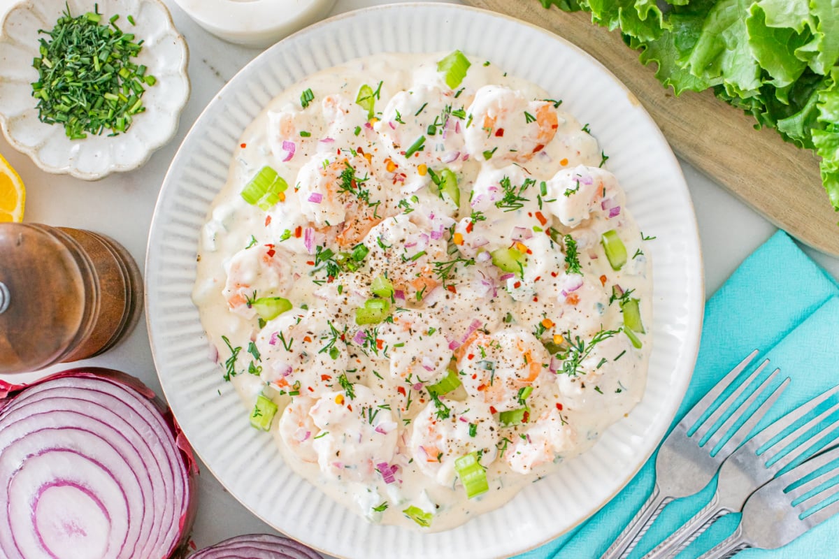 A plate of shrimp salad with creamy dressing, garnished with chopped celery, red onion, herbs, and red pepper flakes, surrounded by fresh ingredients on a table.