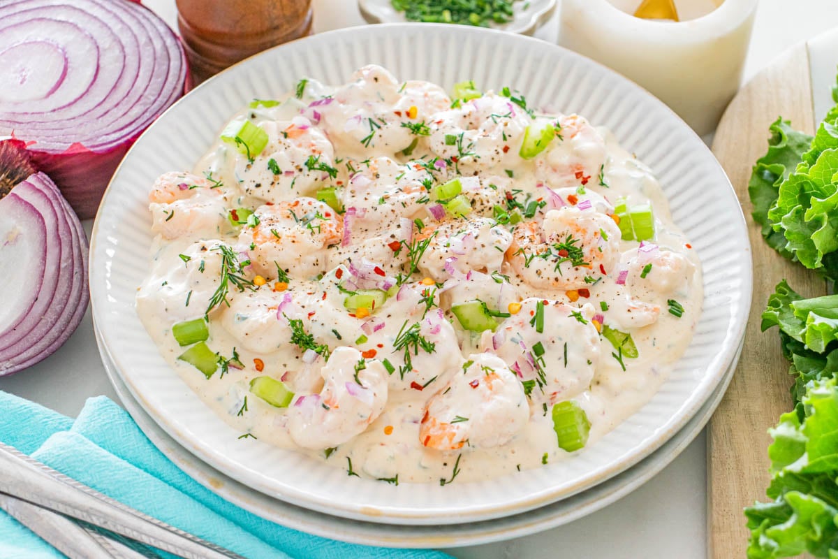 A plate of creamy shrimp salad garnished with chopped celery, red onion, dill, and black pepper, served on a white dish with fresh vegetables nearby.