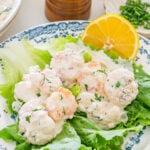 A plate of shrimp salad with creamy dressing, chopped herbs, and red onions served on green lettuce leaves, garnished with a lemon wedge.