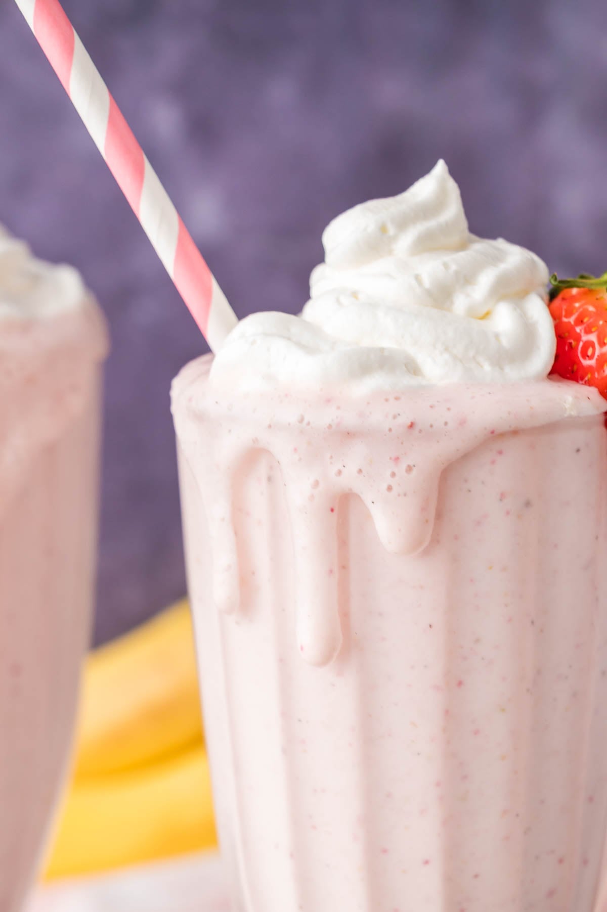 A strawberry milkshake in a glass with whipped cream on top, a pink-striped straw, and a fresh strawberry garnish.