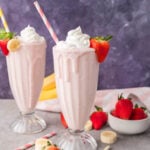 Two tall glasses of strawberry banana milkshake with whipped cream, striped straws, and fresh fruit garnish, set on a gray table with whole strawberries and banana slices nearby.