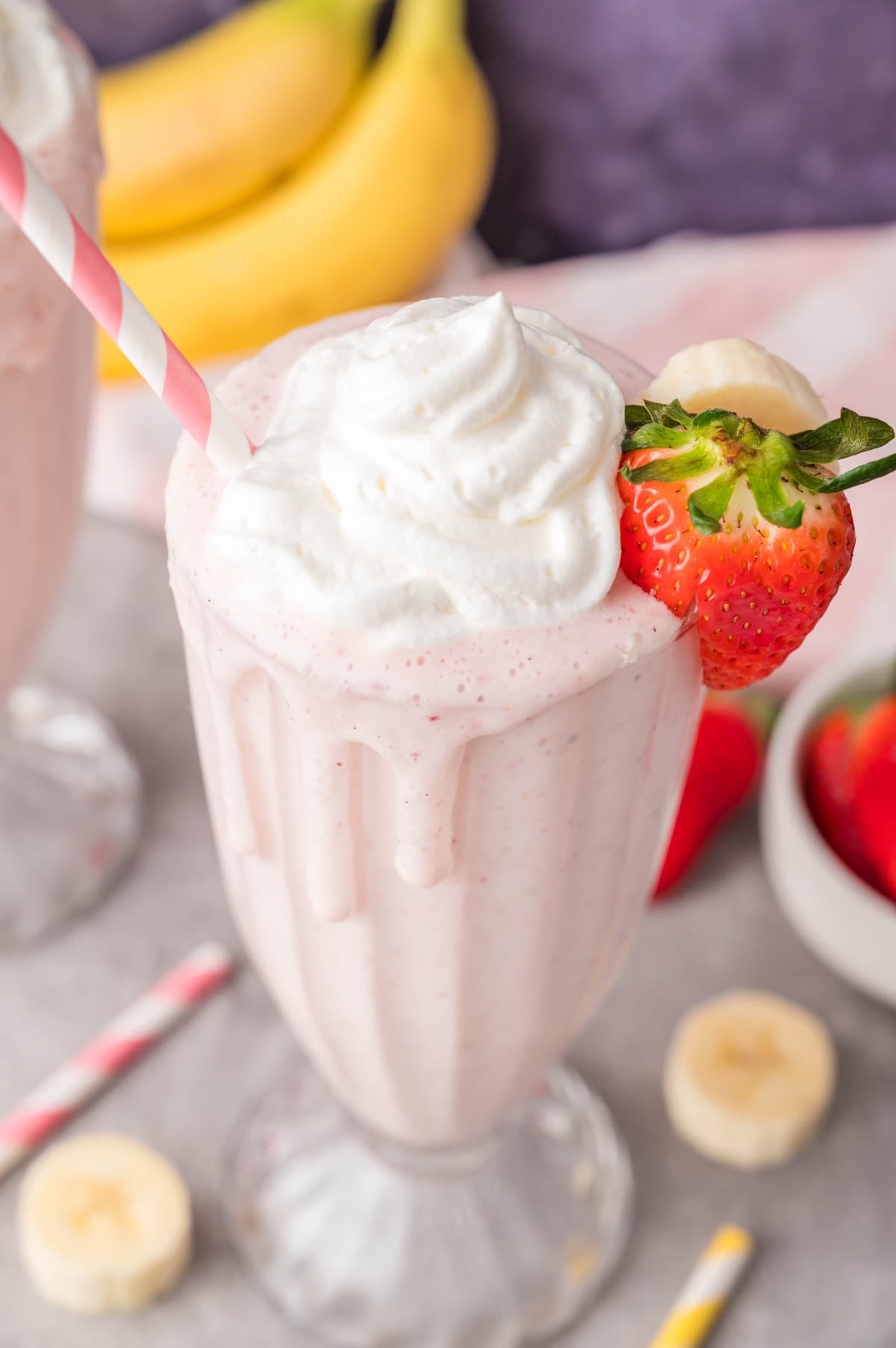 A strawberry banana milkshake topped with whipped cream and garnished with a strawberry and banana slice in a glass with a pink striped straw.