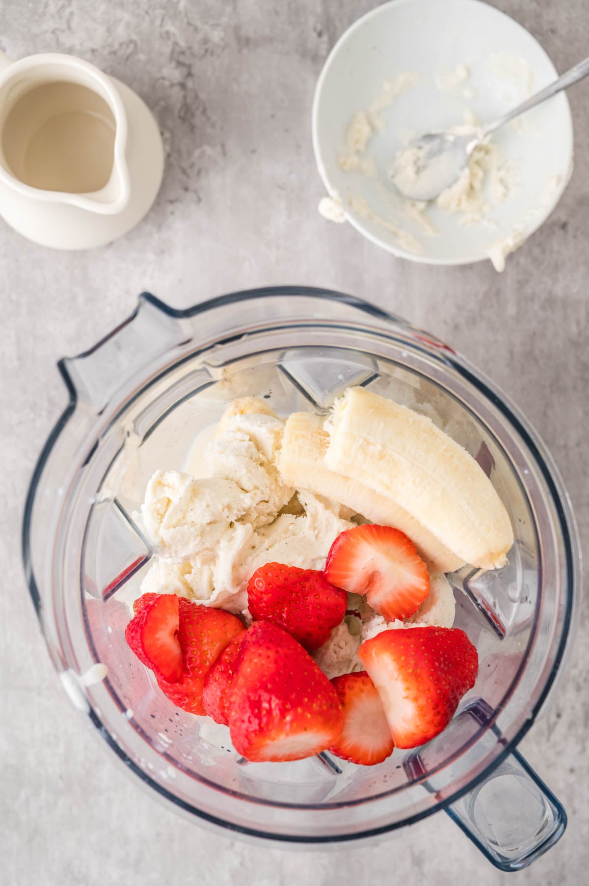 A blender containing vanilla ice cream, a peeled banana, and sliced strawberries sits on a counter near a bowl with a spoon and a small pitcher.