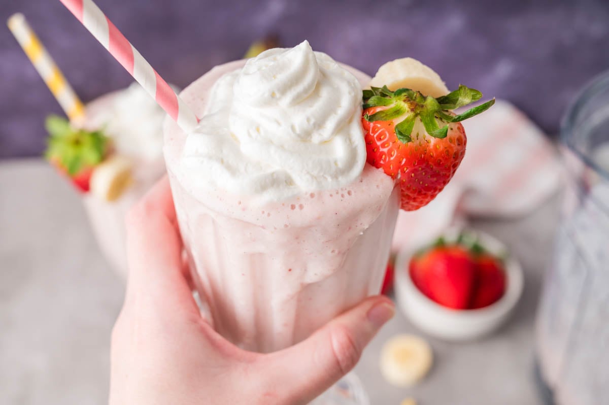 A hand holding a glass of strawberry banana milkshake topped with whipped cream, garnished with a fresh strawberry and banana slice, with a pink striped straw.