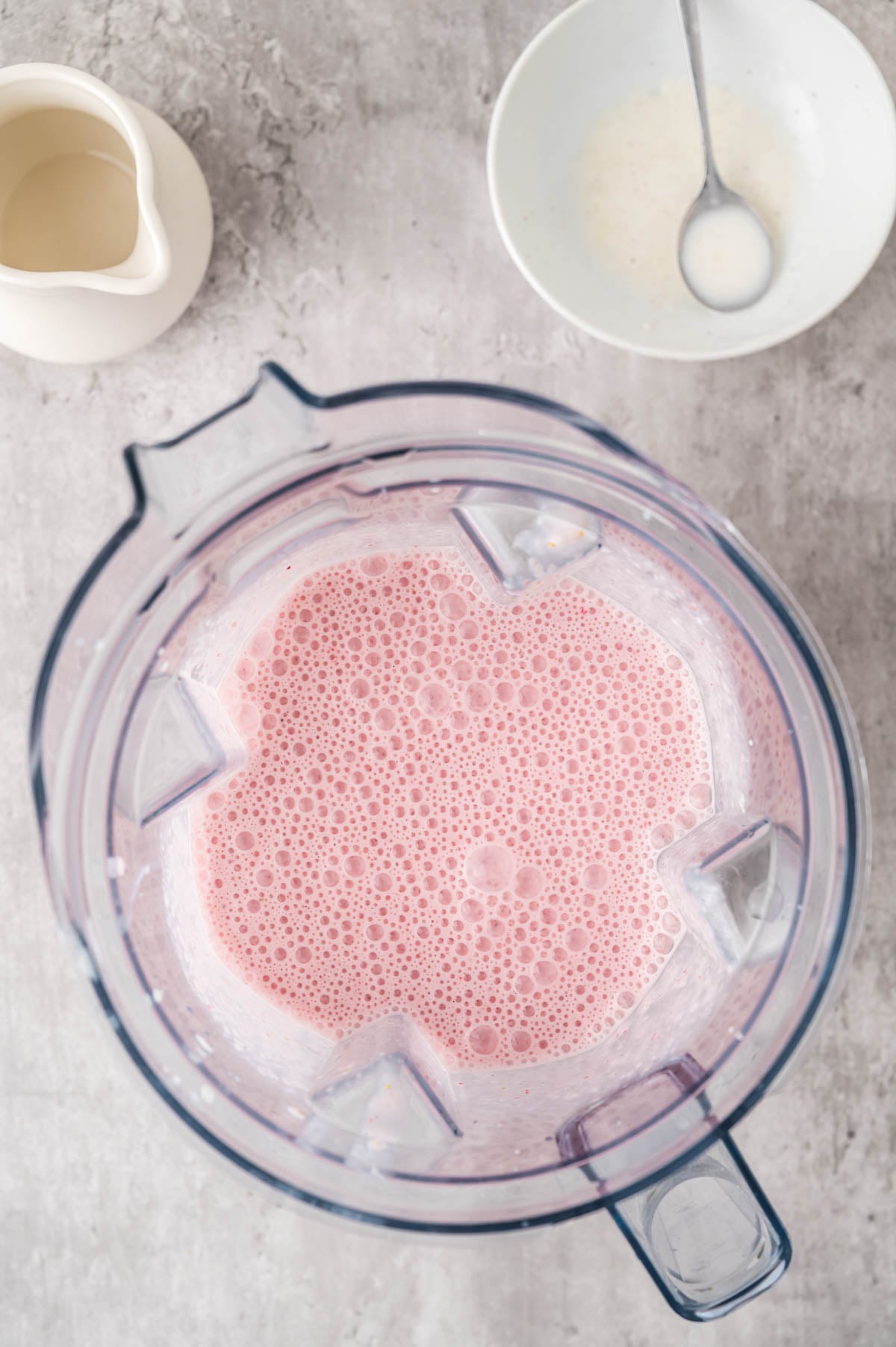 A blender filled with a pink milkshake on a gray surface, next to a white bowl with a spoon and a small white pitcher.