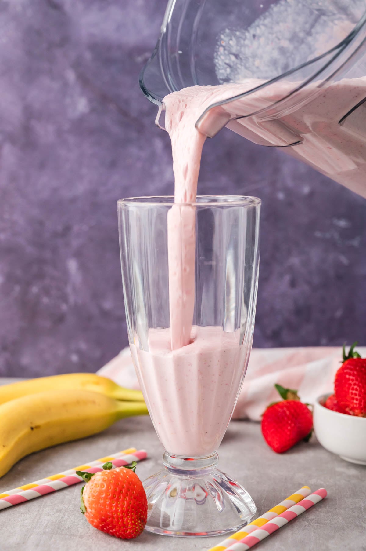 A thick pink milkshake is being poured from a blender into a tall glass, with strawberries, bananas, and colorful straws nearby on a gray surface.