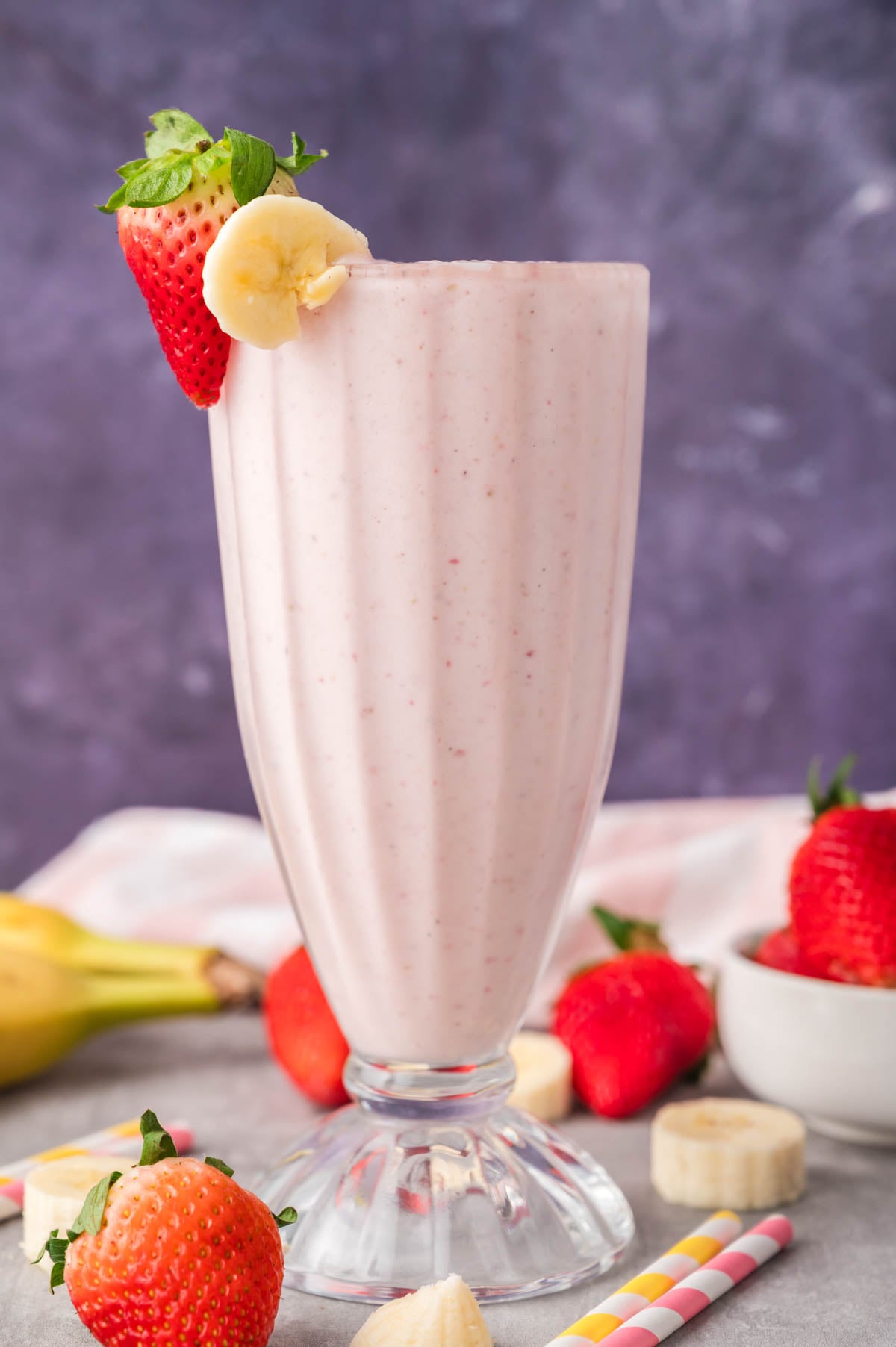 A tall glass of strawberry banana milkshake garnished with a strawberry and banana slice, surrounded by fresh strawberries and bananas, with two striped straws nearby.