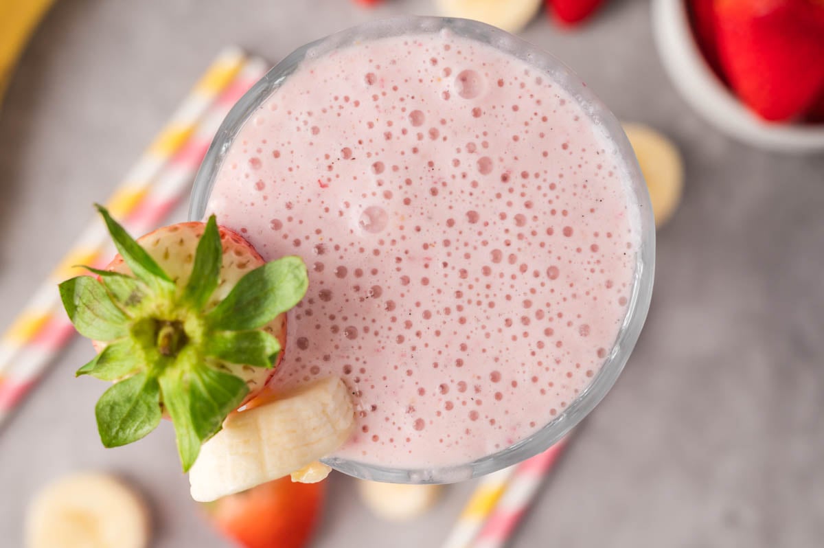 A glass of pink milkshake topped with a whole strawberry and banana slice, with straws and fruit pieces visible in the blurred background.