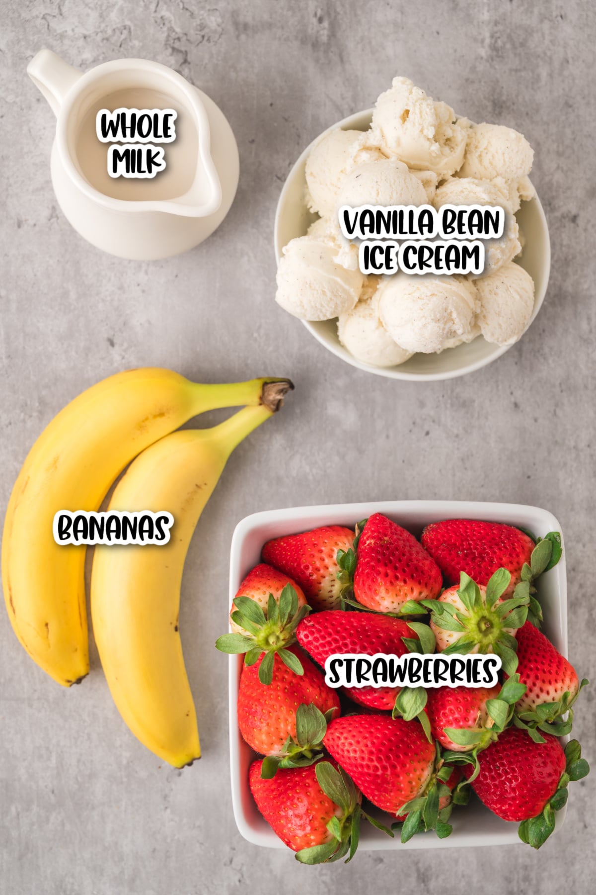 Top-down image of whole milk in a jug, a bowl of vanilla bean ice cream, two bananas, and a container of fresh strawberries on a gray surface.