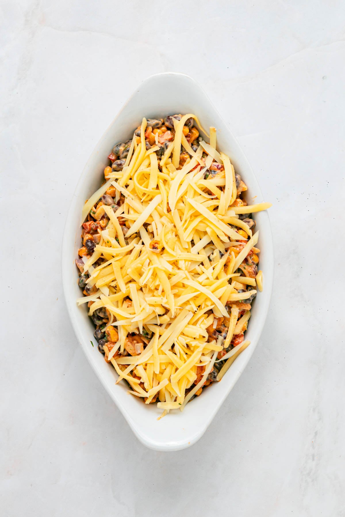 An oval white baking dish filled with a mixture of vegetables, topped with a layer of shredded cheese.