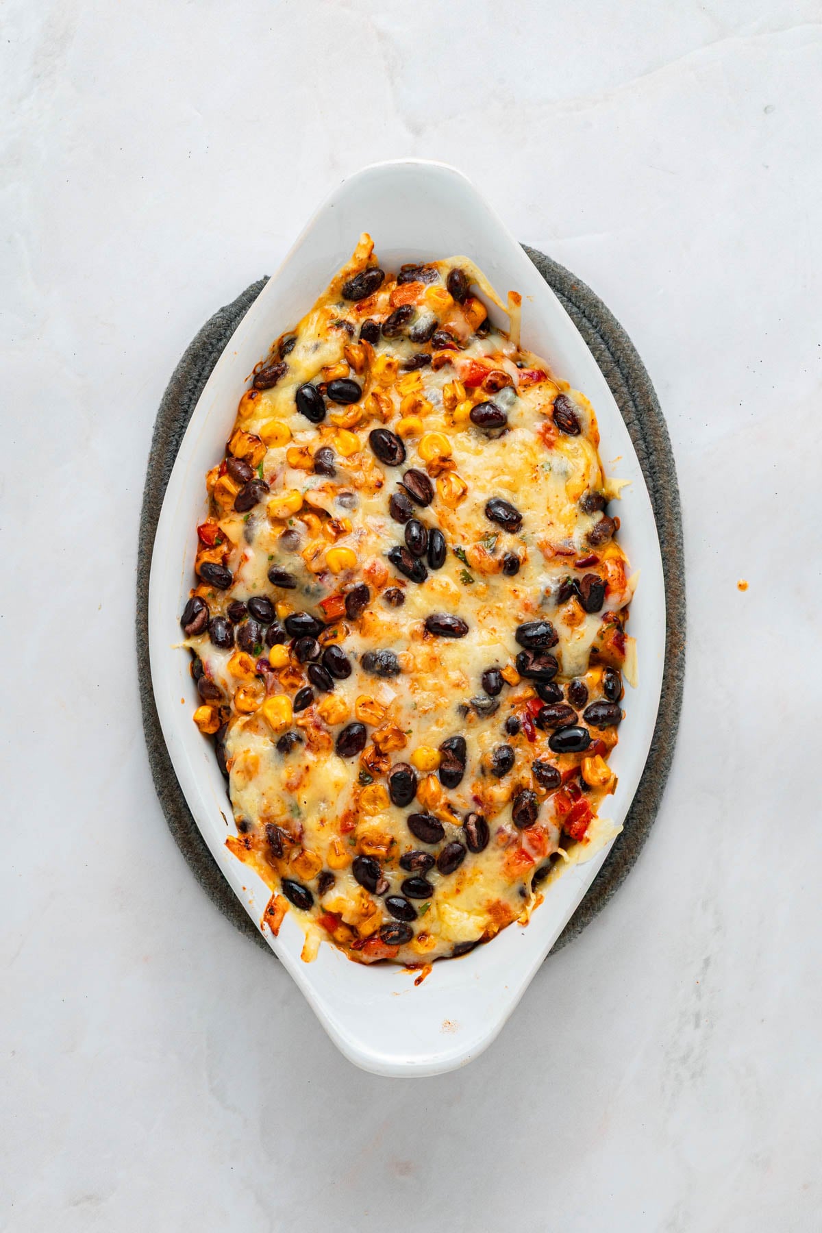 Oval baking dish filled with a baked casserole topped with melted cheese, black beans, corn, and vegetables, placed on a dark grey trivet.
