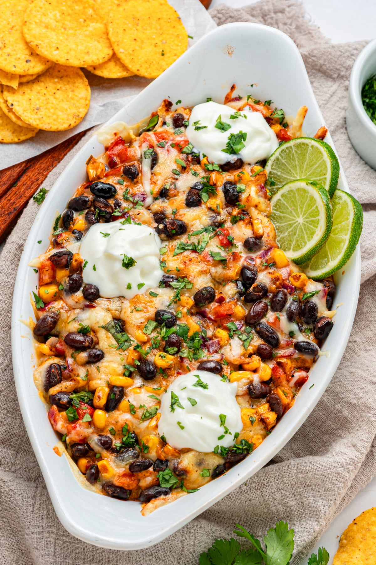 A baked black bean and corn dip topped with melted cheese, sour cream, lime slices, and chopped herbs, served next to tortilla chips.