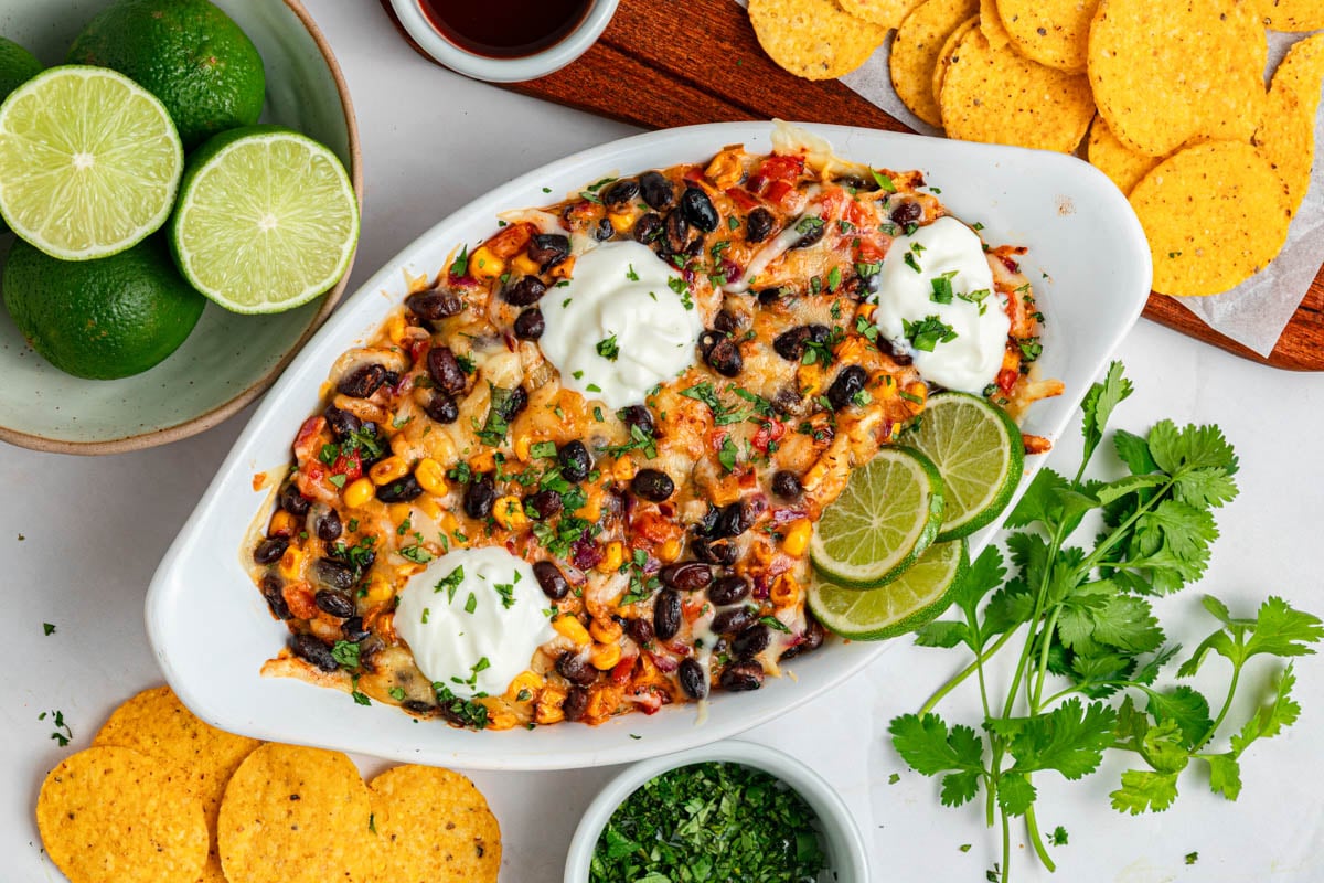 A baked Mexican-inspired dip topped with melted cheese, black beans, corn, sour cream, lime slices, and chopped herbs, surrounded by tortilla chips, fresh limes, and cilantro.