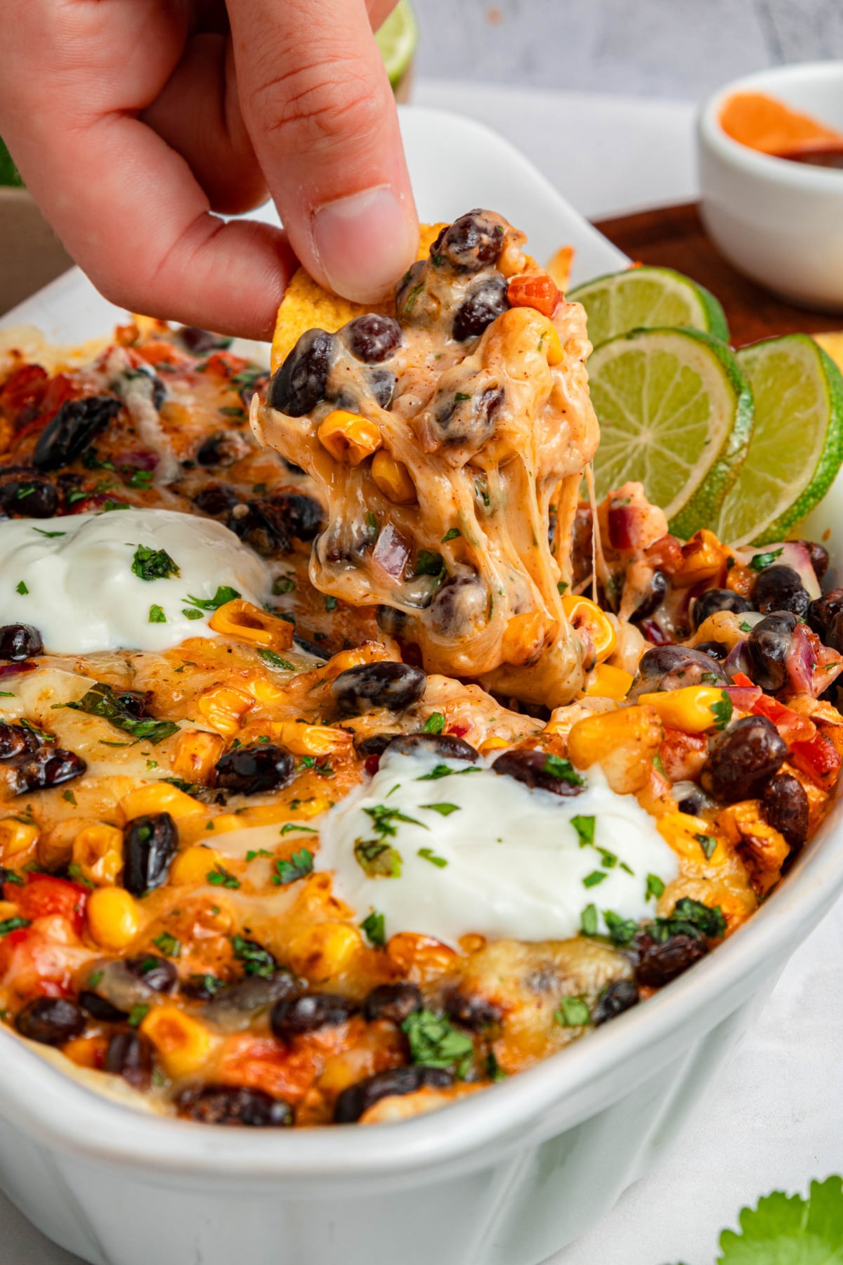A hand dips a tortilla chip into a baked dip topped with melted cheese, black beans, corn, and sour cream, garnished with chopped herbs and lime slices.