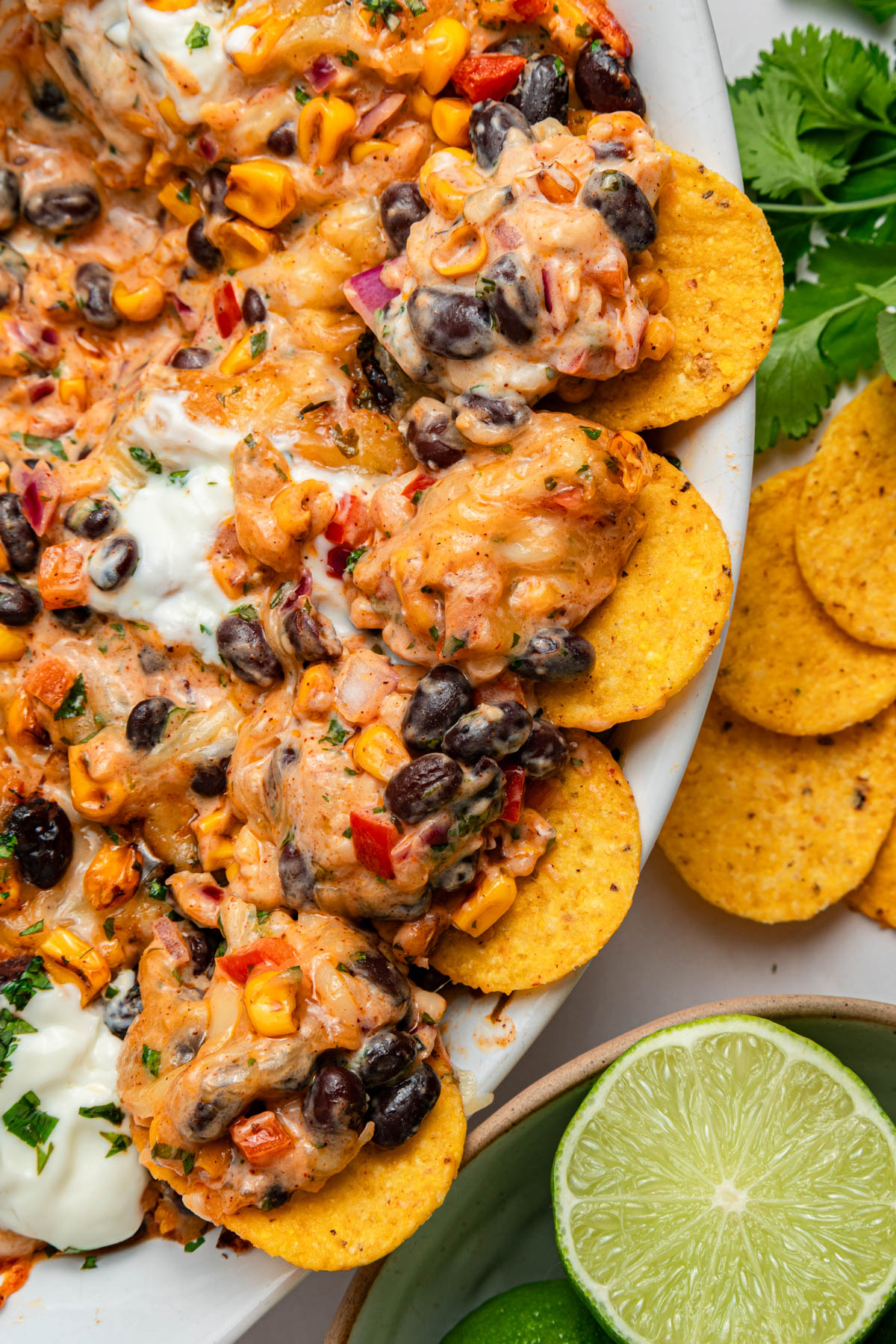 A close-up of cheesy baked dip with black beans, corn, red peppers, and tortilla chips, garnished with herbs. Lime halves and fresh cilantro are on the side.