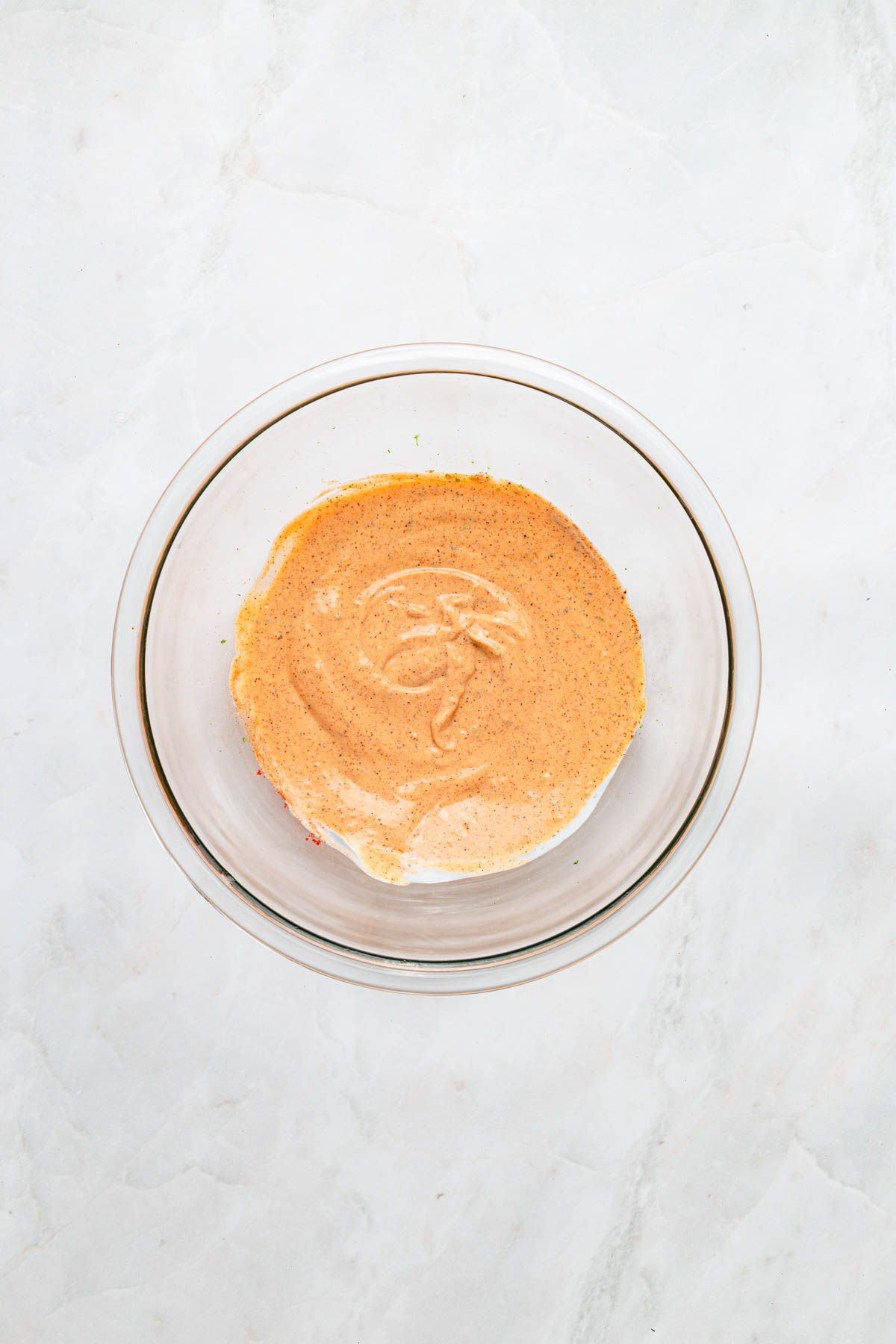 A glass bowl filled with a light brown, creamy sauce or dressing sits on a white marble surface.