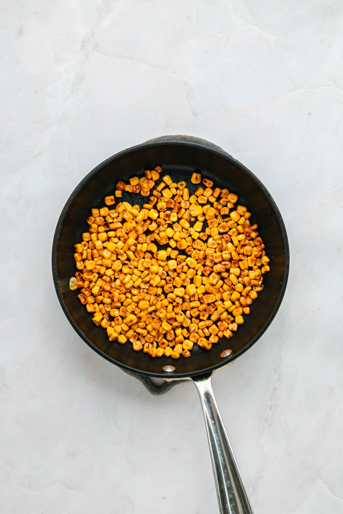 A frying pan containing cooked, browned corn kernels sits on a light-colored surface.
