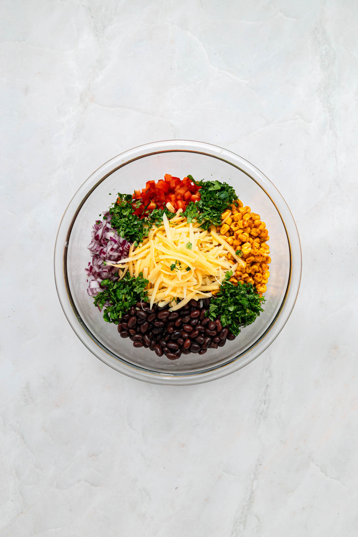 A glass bowl containing chopped red onion, red bell pepper, cilantro, shredded cheese, black beans, corn, and parsley, arranged in separate sections on a light surface.