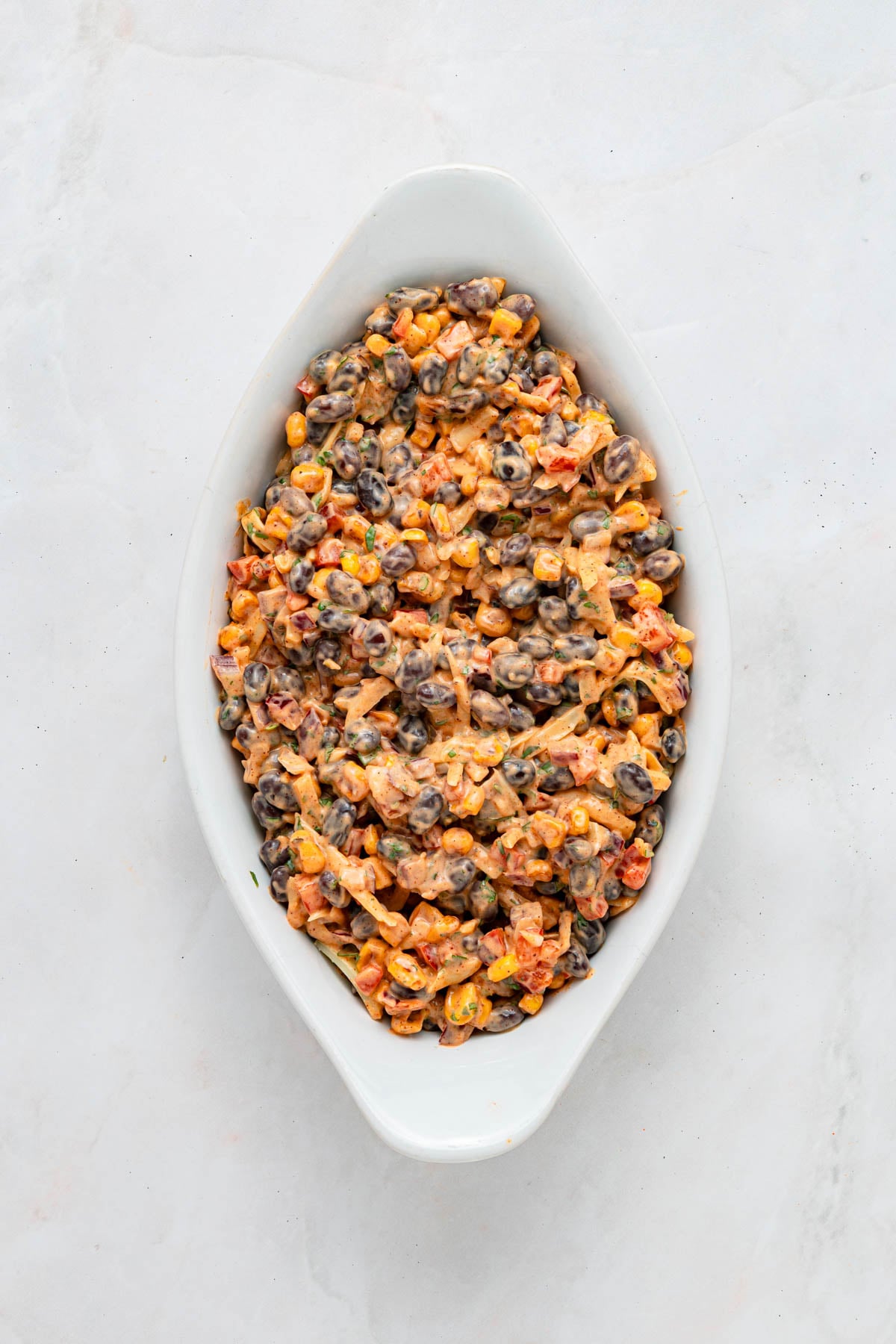 An oval white dish filled with a mixture of black beans, corn, diced tomatoes, and melted cheese on a light surface.