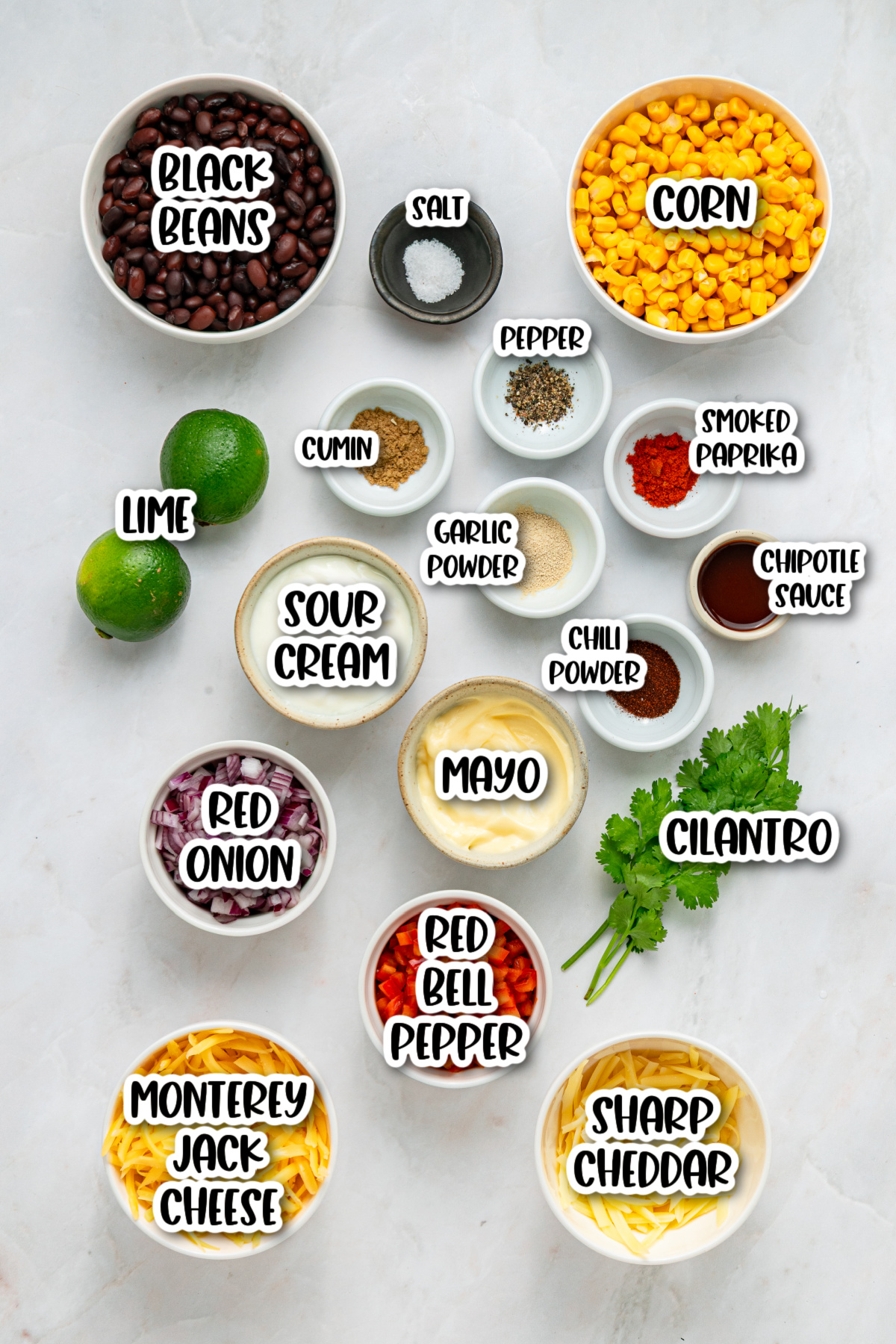 A top-down view of bowls containing black beans, corn, red onion, red bell pepper, seasonings, lime, mayo, sour cream, chipotle sauce, cilantro, and shredded cheeses on a white surface.
