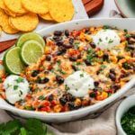A baked Mexican-style dip with black beans, corn, cheese, and herbs, topped with sour cream and lime slices. Tortilla chips are served on the side.