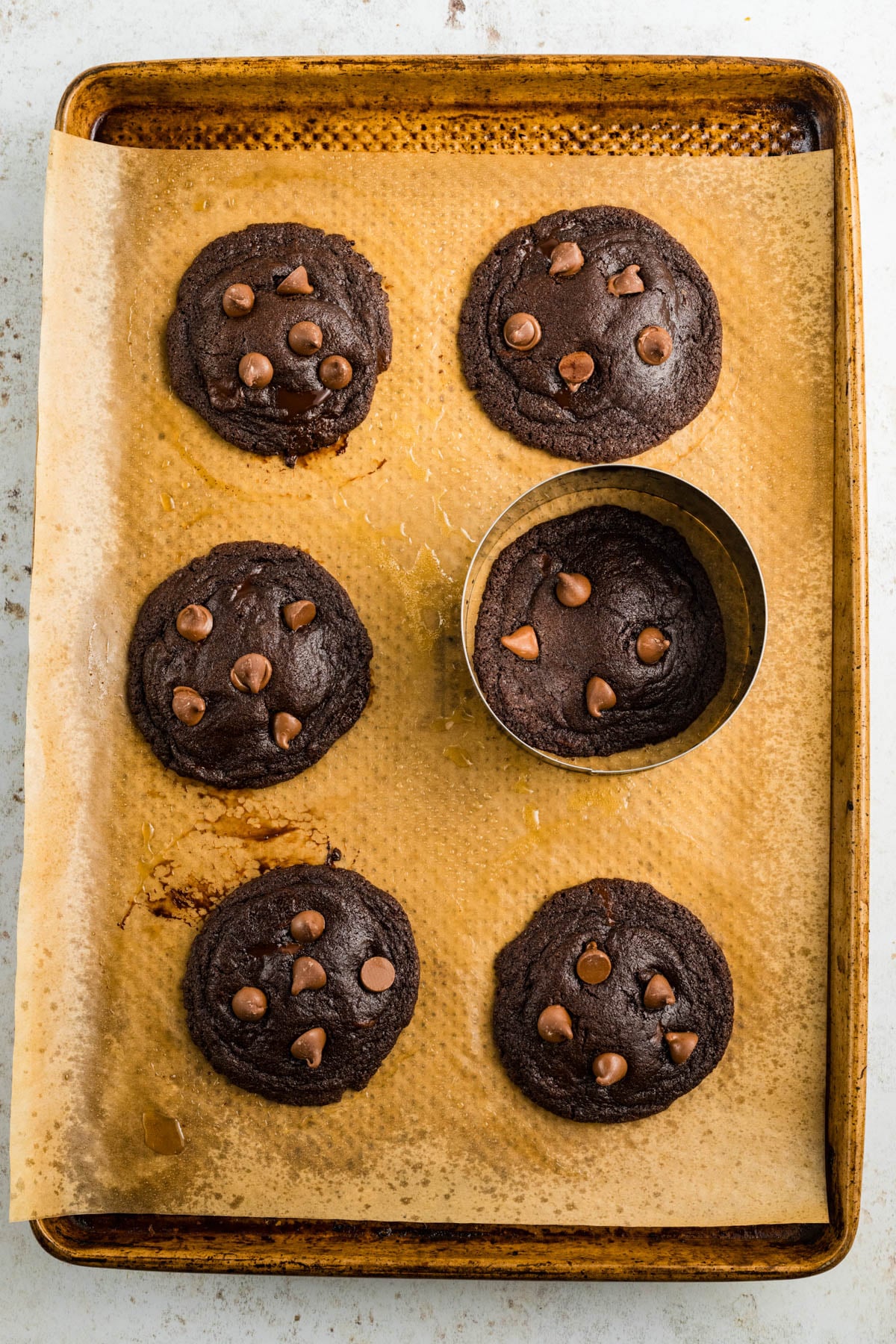 Six chocolate chocolate chunk cookies on parchment paper atop a baking sheet; a round cookie cutter circles one of the cookies.