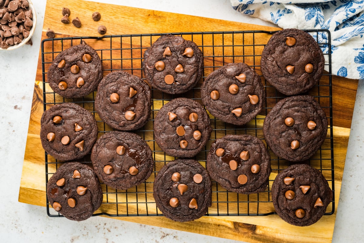 Chocolate chocolate chunk cookies cooling on a black wire rack placed on a wooden cutting board.