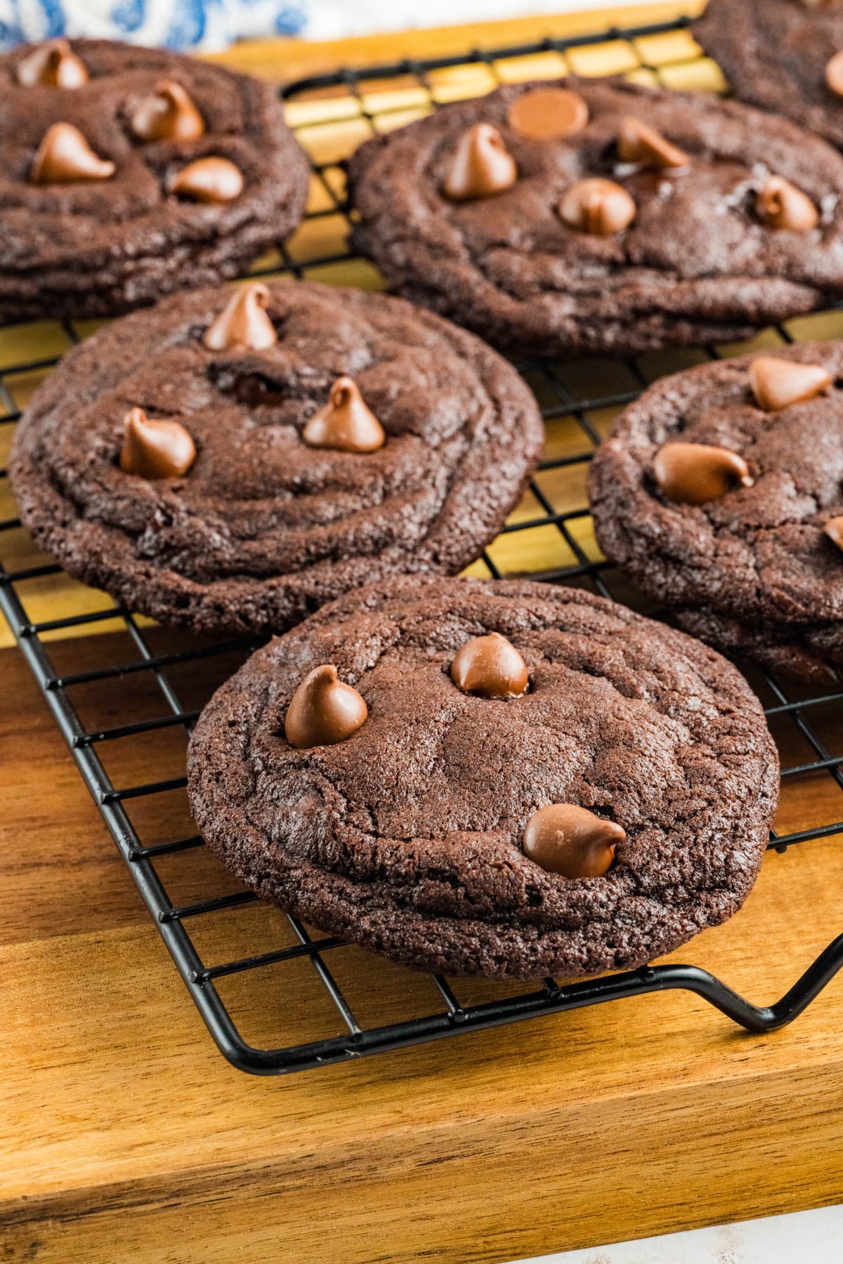 Chocolate chocolate chunk cookies with chocolate chips cooling on a black wire rack placed on a wooden surface.