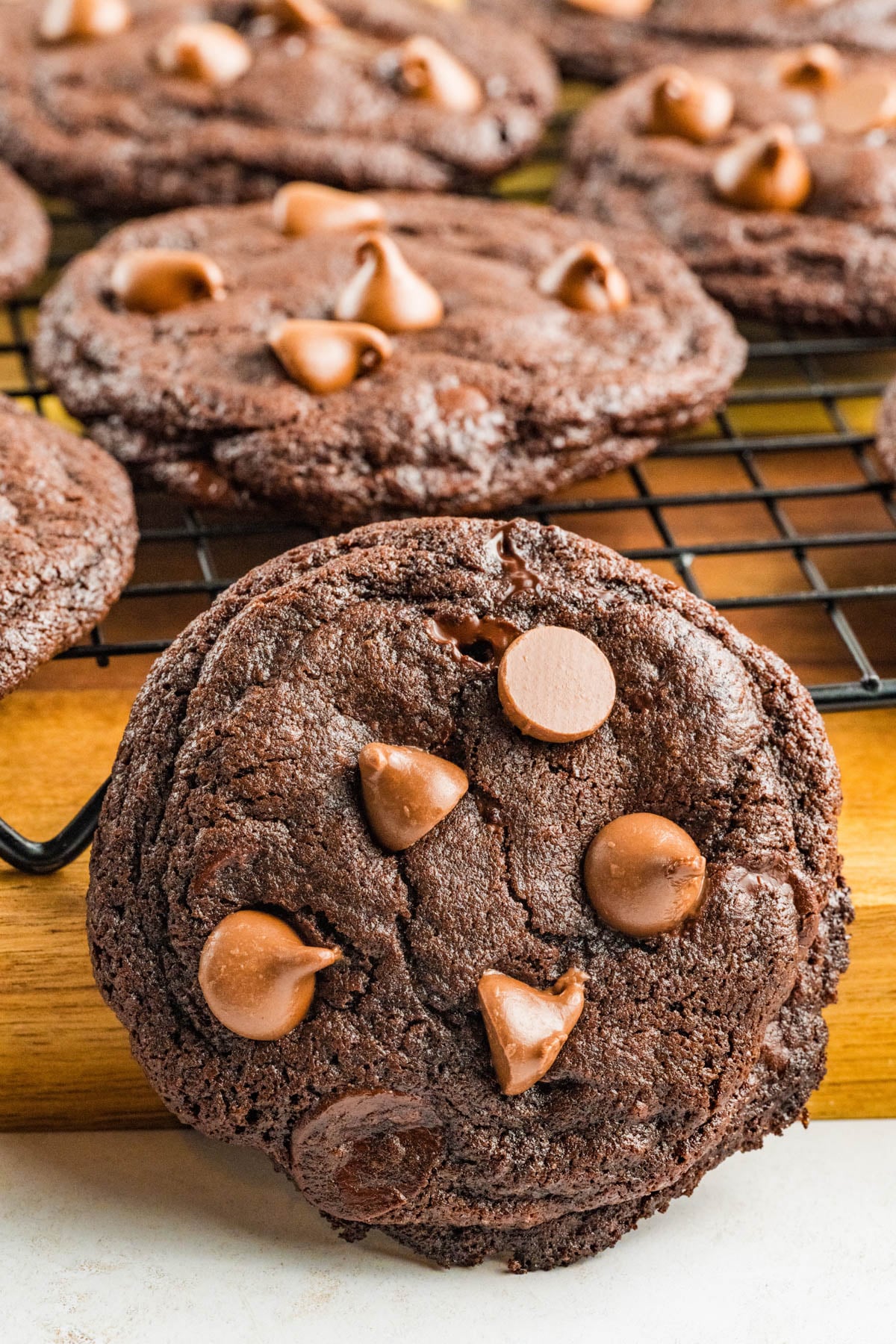 A close-up of chocolate chocolate chunk cookies, some resting on a cooling rack and one in the foreground on a wooden surface.