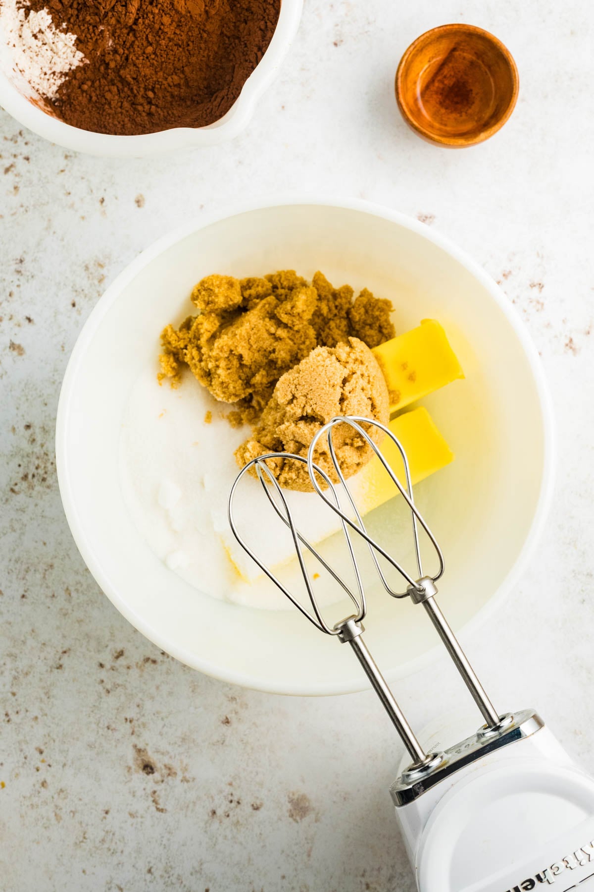 A mixing bowl with brown sugar, white sugar, and butter next to an electric hand mixer; cocoa powder and a small bowl are seen nearby on a white countertop.