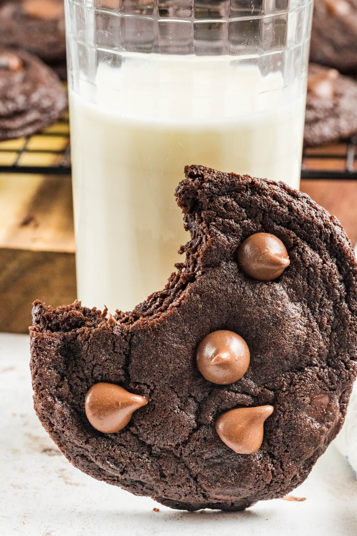 A bitten chocolate cookie with chocolate chips rests in front of a glass of milk, with more cookies cooling on a rack in the background.